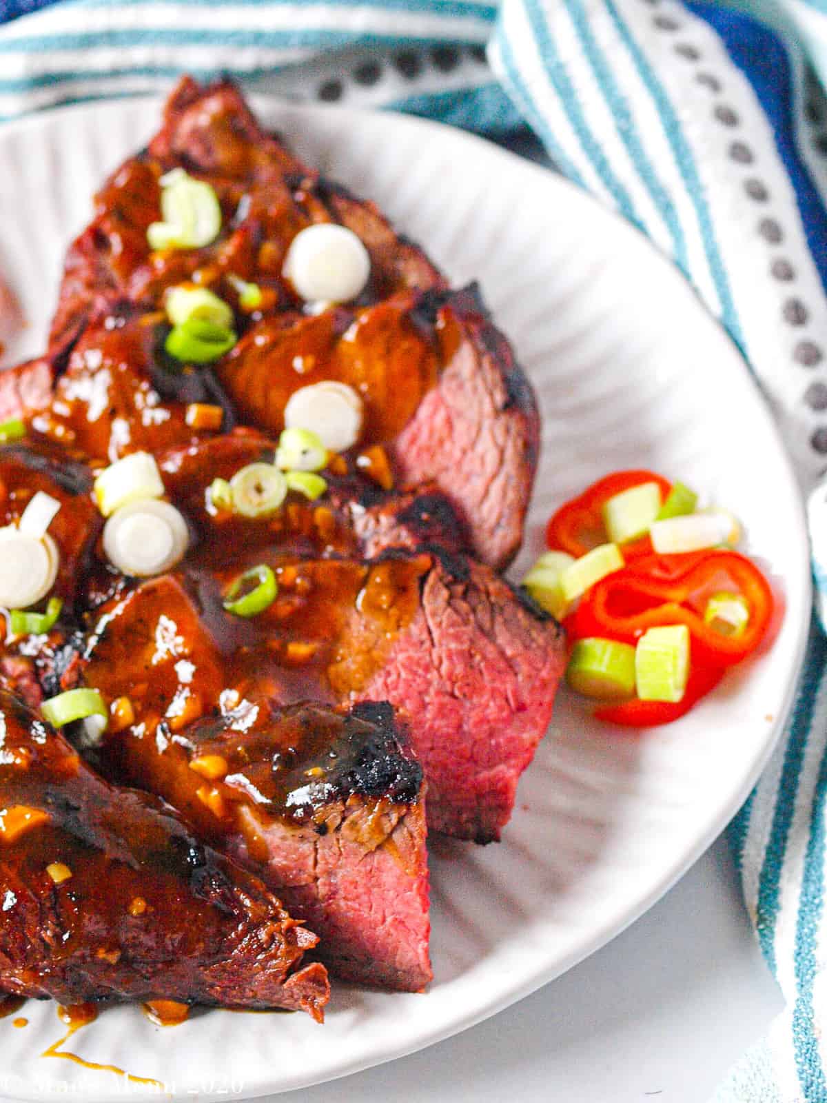 An up close of pieces of grilled tri tip with a sweet soy glaze, green onions, and red peppers on top. 