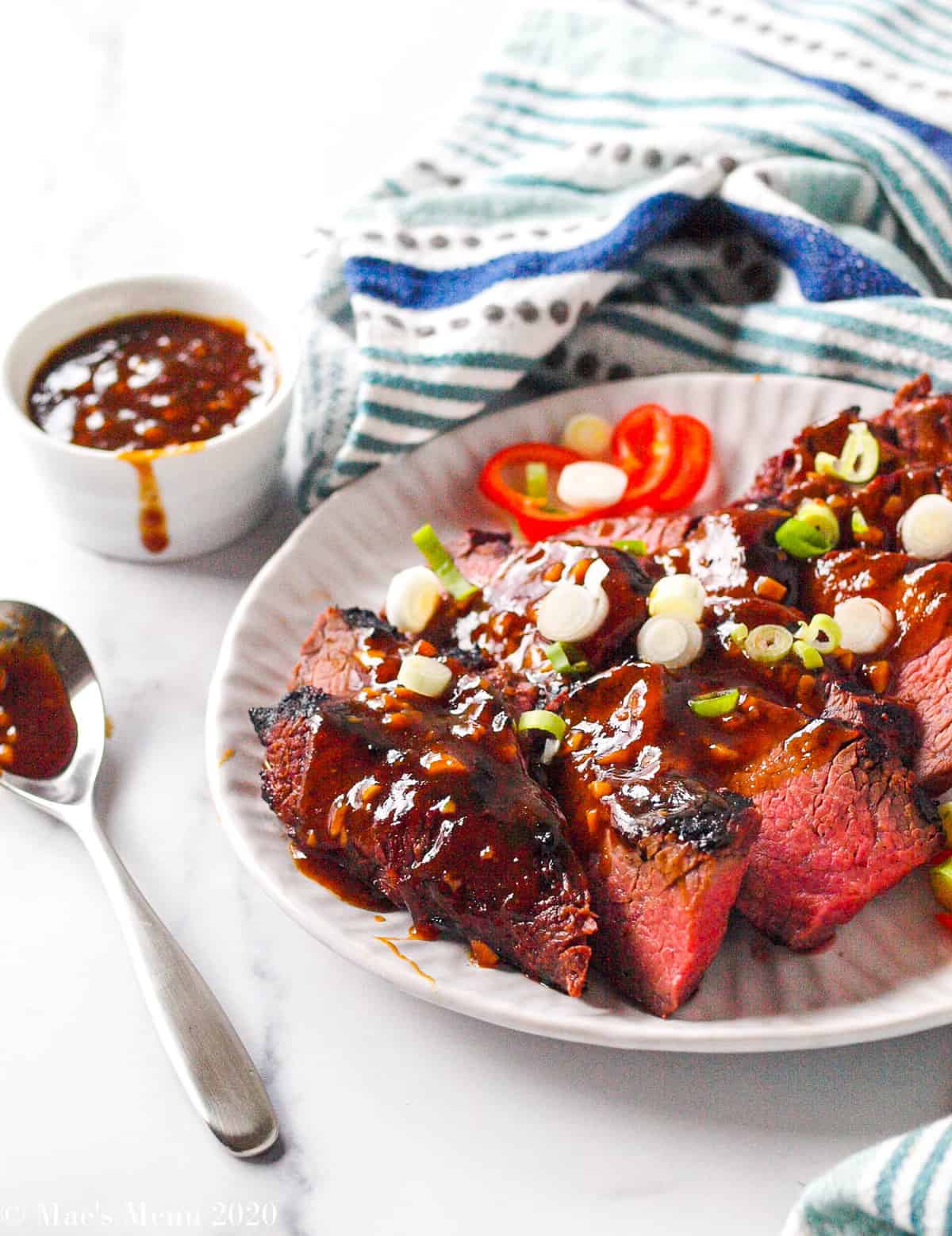 a plate of sliced trip tip with sweet soy sauce glaze. Beside the plate sits a spoonful of glaze and a cup of glaze.