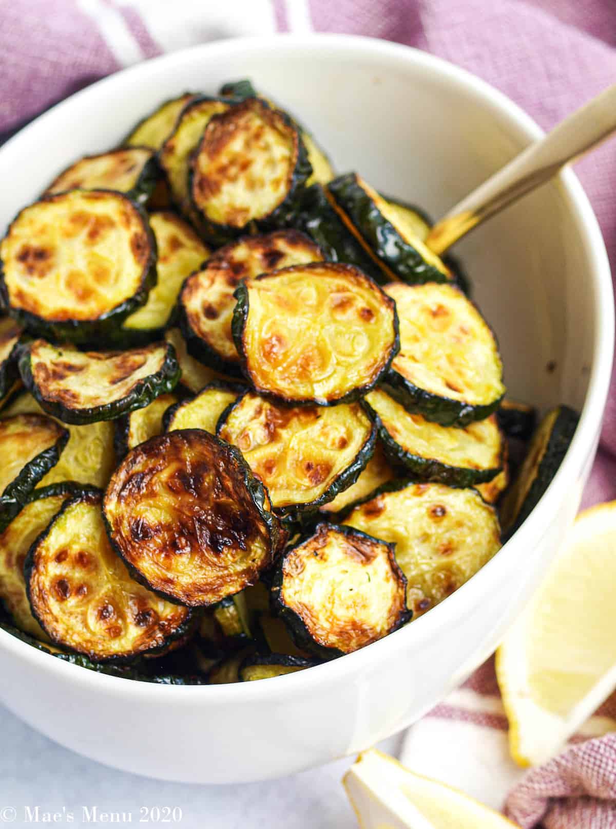 A picture of a bowl of air fryer zucchini by lemon slices and a purple towel
