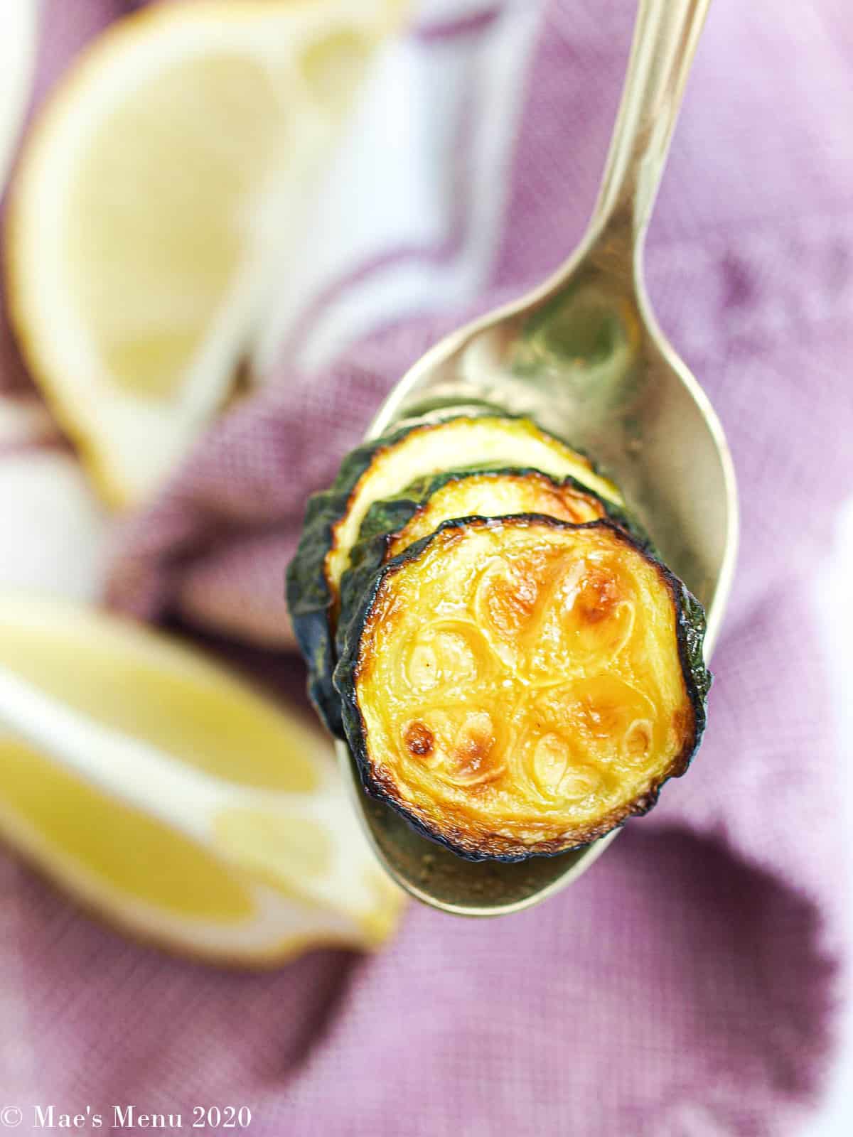 A gold spoon with air fryer zucchini on it. In the background is a purple towel and lemon slicesThis Air Fryer Zucchini is one of my favorite zucchini recipes! They made a delicious low carb side dish and are super tasty! They're one of my go-to air fryer recipes, especially when the zucchini is fresh and sweet! Make this healthy side dish for dinner this week! #zucchini #zucchinirecipes #healthysidedish #airfryer #airfryervegetables #airfryerzucchini