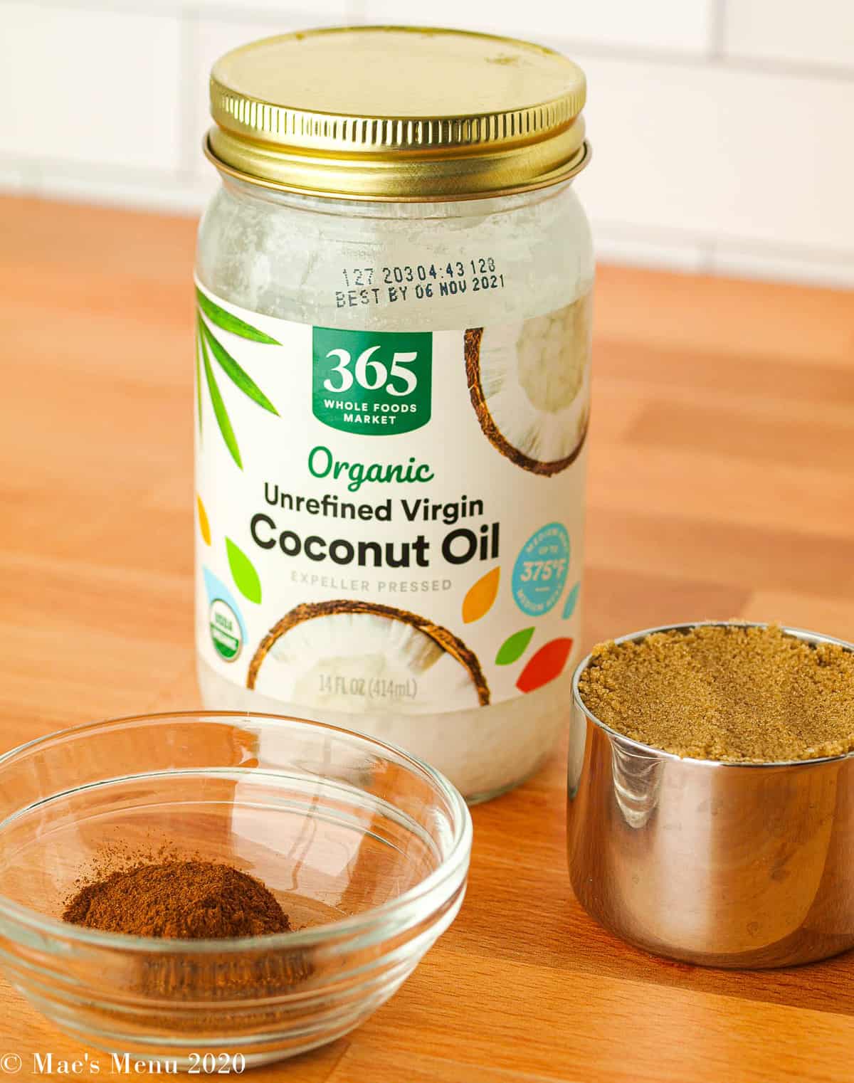 A jar of coconut oil, small measuring cup of dark brown sugar, and a small clear dish of cinnamon and spices.