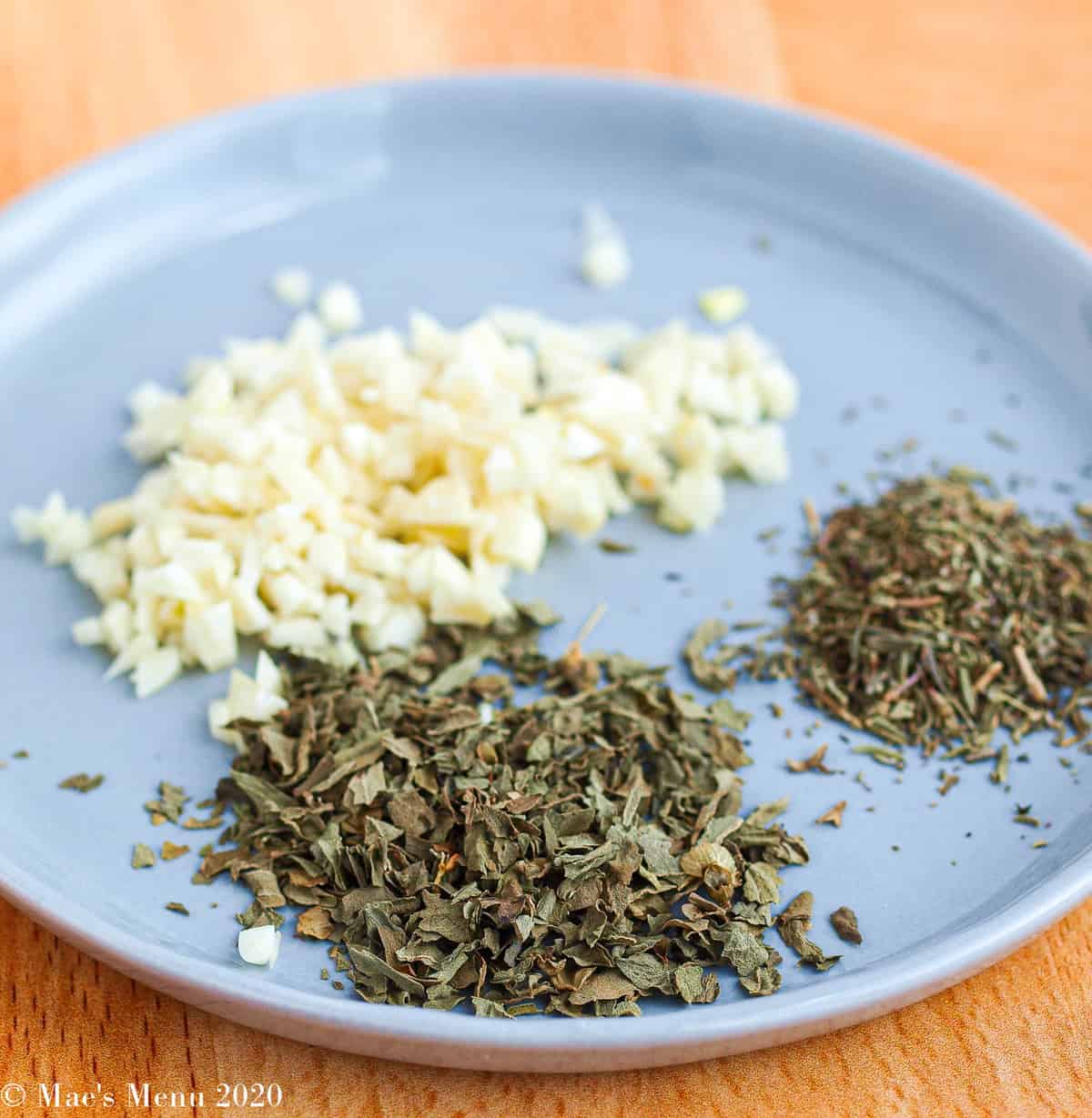 A small blue plate with piles of minced garlic, dried basil, and dried thyme on it.