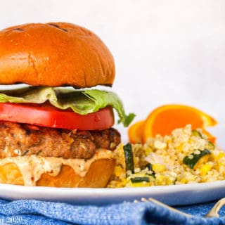 A large Jamaican jerk turkey burger on a white plate next to quinoa salad and oranges.