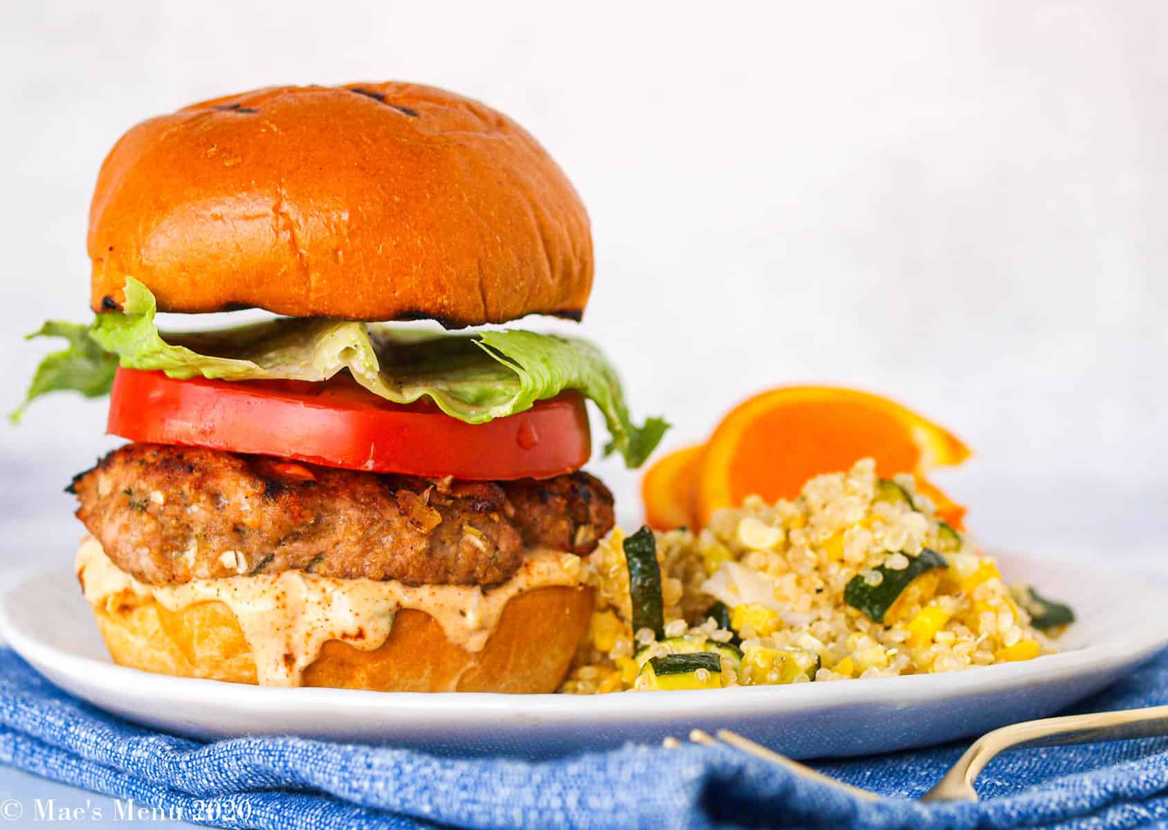 A large Jamaican jerk turkey burger on a white plate next to quinoa salad and oranges.