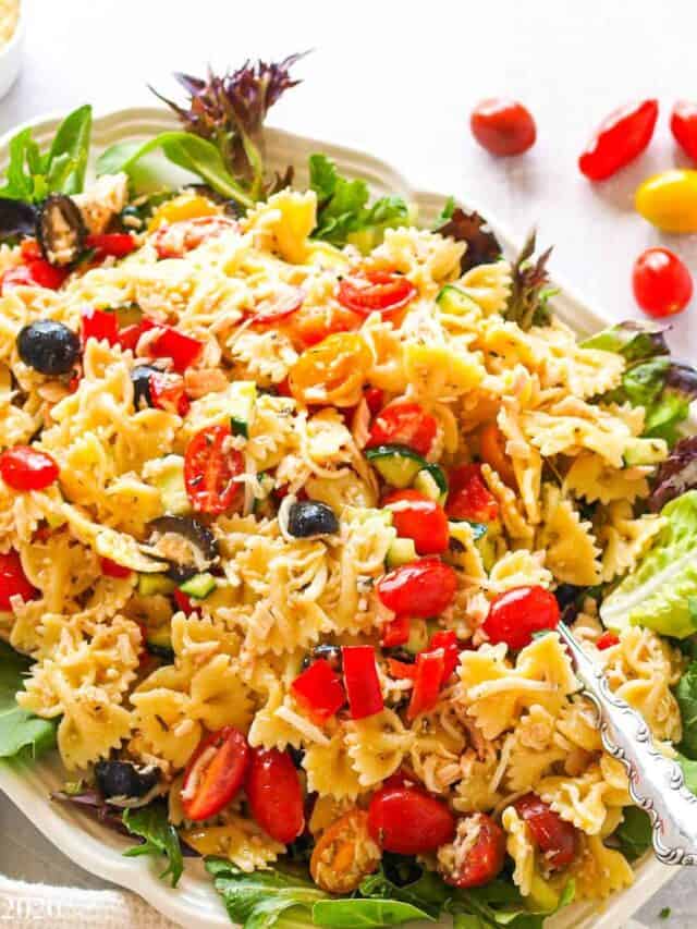 An overhead shot of a large platter of Italian Tuna Pasta salad with tomatoes, a glass of water, and a cup of cheese next to the platter.