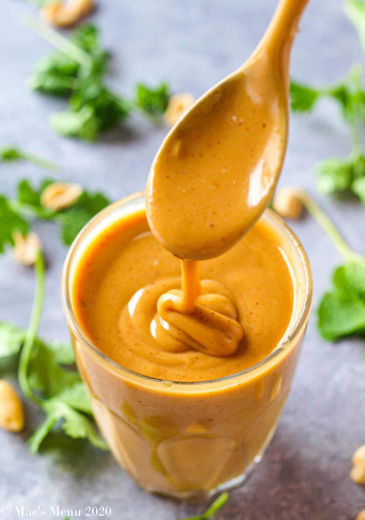 A side angle shot of a cup of peanut dressing with a spoon drizzling the dressing into it