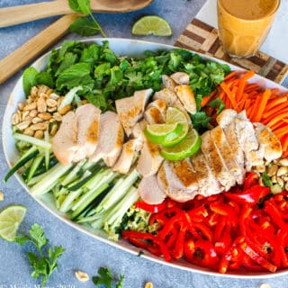 An overhead shot of a platter of thai chicken salad with peanut dressing. Besides the platter sits the cup of dressing. To the top of the platter sits the salad tongs. Chopped herbs, lime, and peanuts sit around the platter