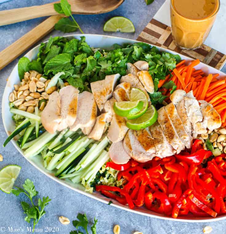 An overhead shot of a platter of thai chicken salad with peanut dressing. Besides the platter sits the cup of dressing. To the top of the platter sits the salad tongs. Chopped herbs, lime, and peanuts sit around the platter
