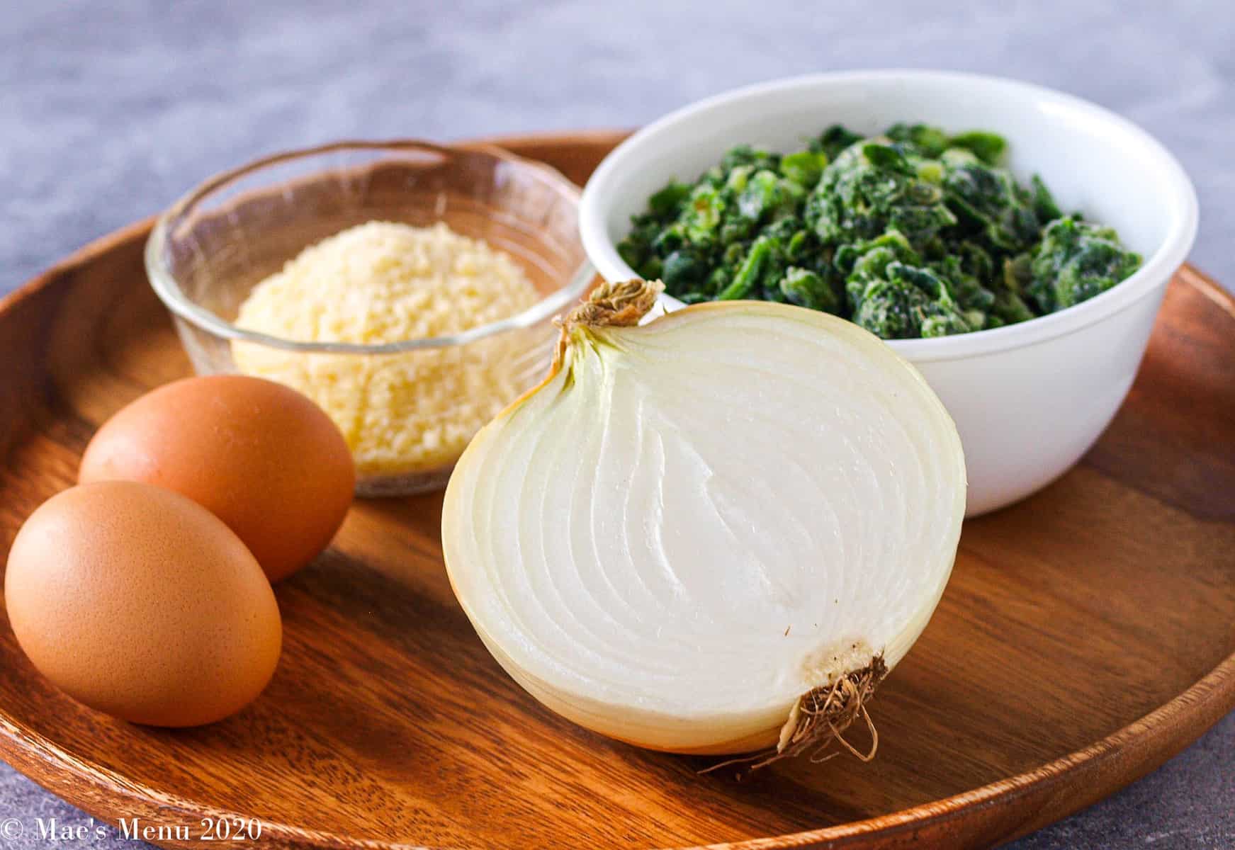 A side angle shot of a wooden tray of ingredients: eggs, parmesan cheese, an onion, and frozen spinach