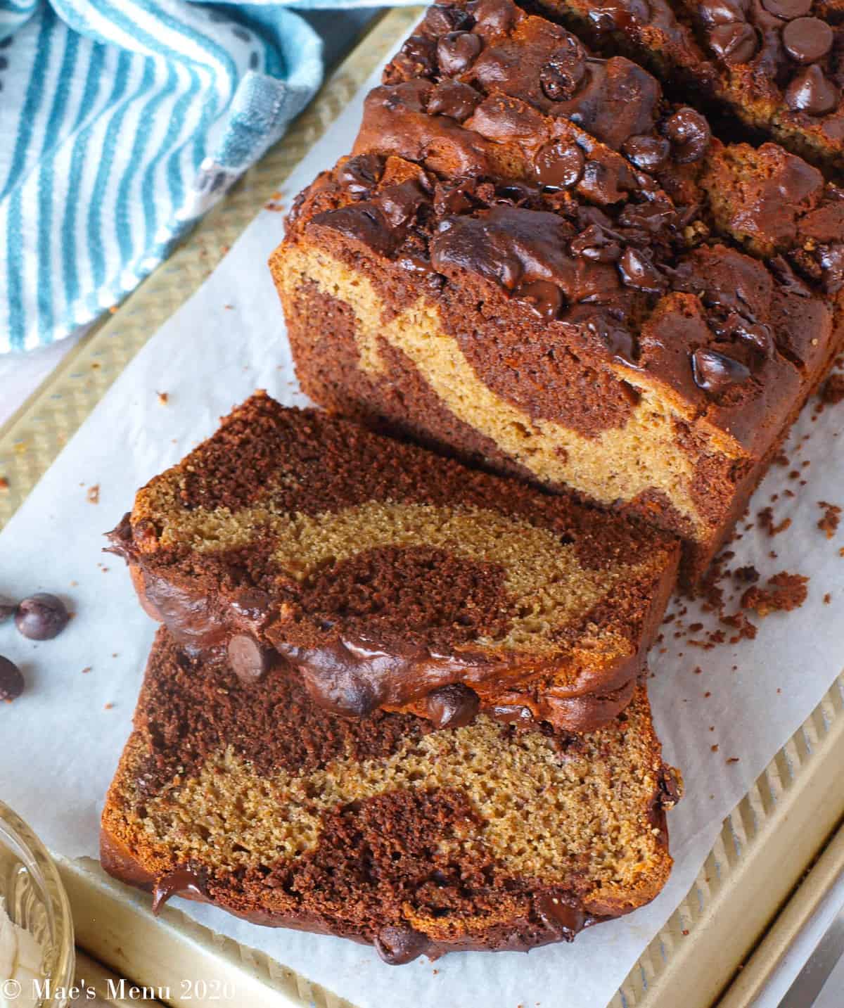 An overhead shot of a loaf of chocolate peanut butter banana bread sliced on a golden pan with chocolate chips and a blue striped towel