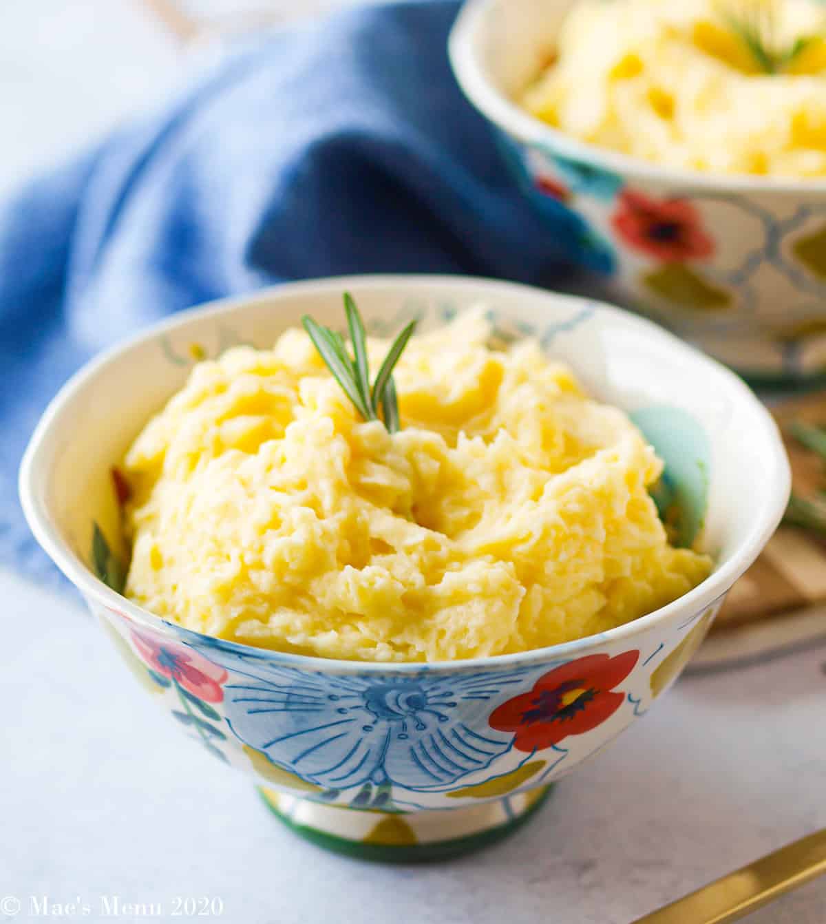 A side angle shot of two bowls of healthy mashed potatoes with sprigs of rosemary on top. In the backround is a blue tea towel