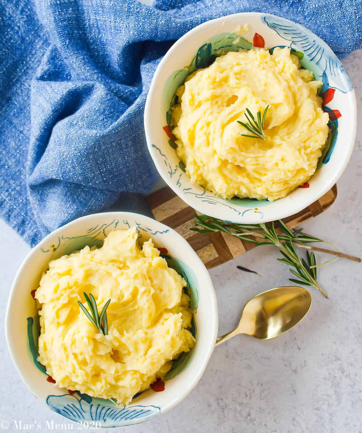 An overhead shot of two bowls of mashed potatoes without butter with a sprig of rosemary and a golden spoon between them