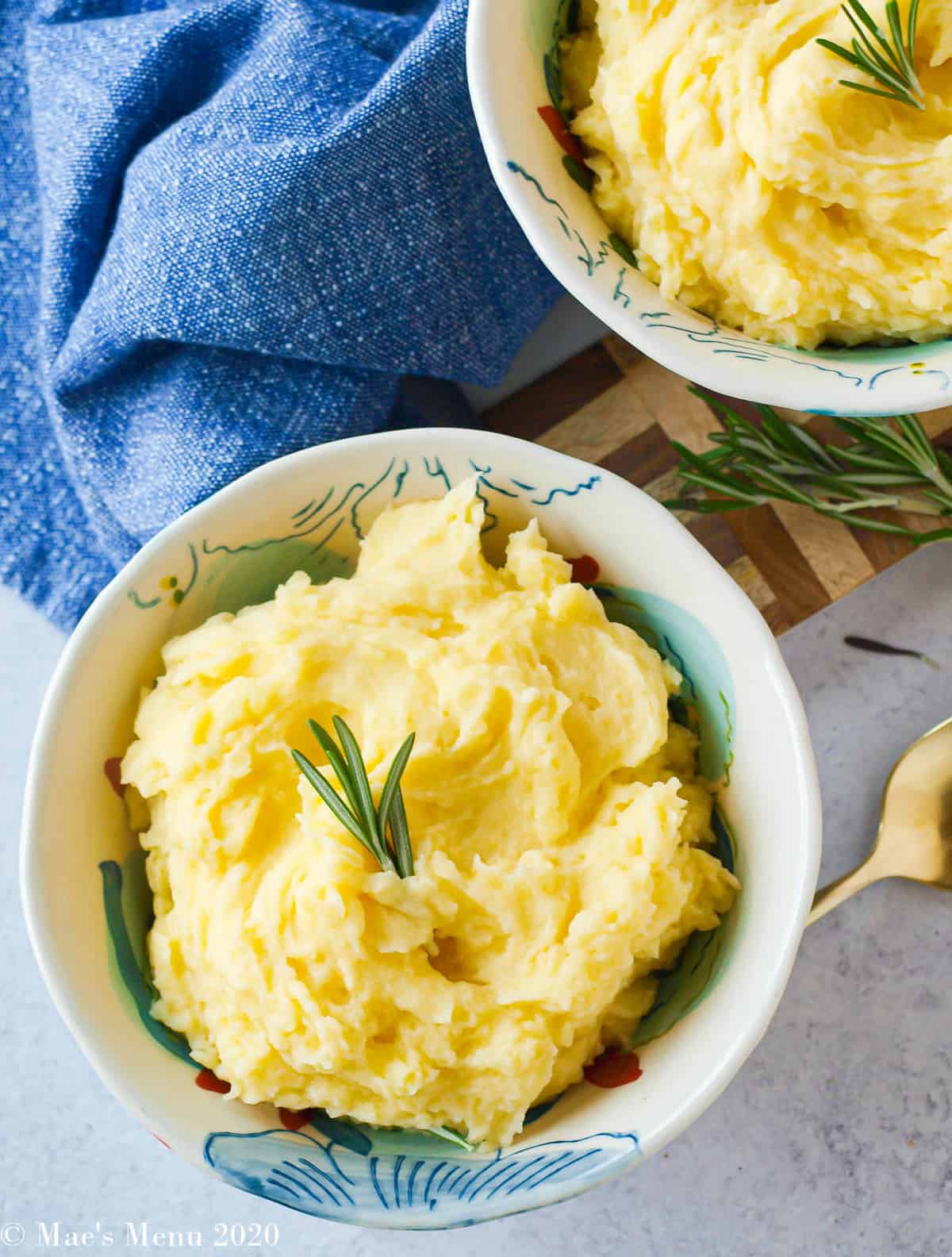 AN overhead shot of two bowls of healthy mashed potatoes without butter with rosemary sprigs on top and a gold spoon between the bowls