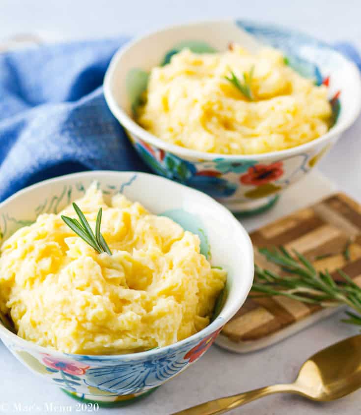 A side angle shot of two bowls of healthy mashed potatoes without butter without oil. A blue tea towel sits in the background and the gold spoon sits in the foreground with a sprig of rosemary and a wooden cutting board