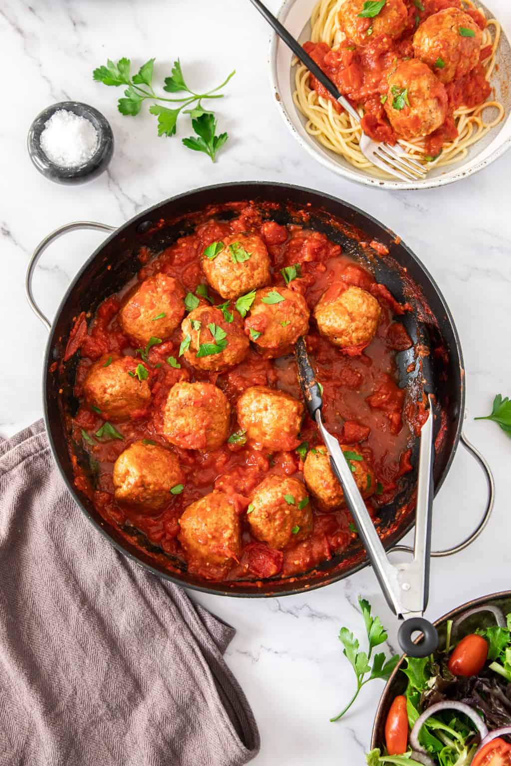 An overhead shot of a large black skillet of turkey meatballs. Next to the skillet is a small white bowl of pasta with meatballs