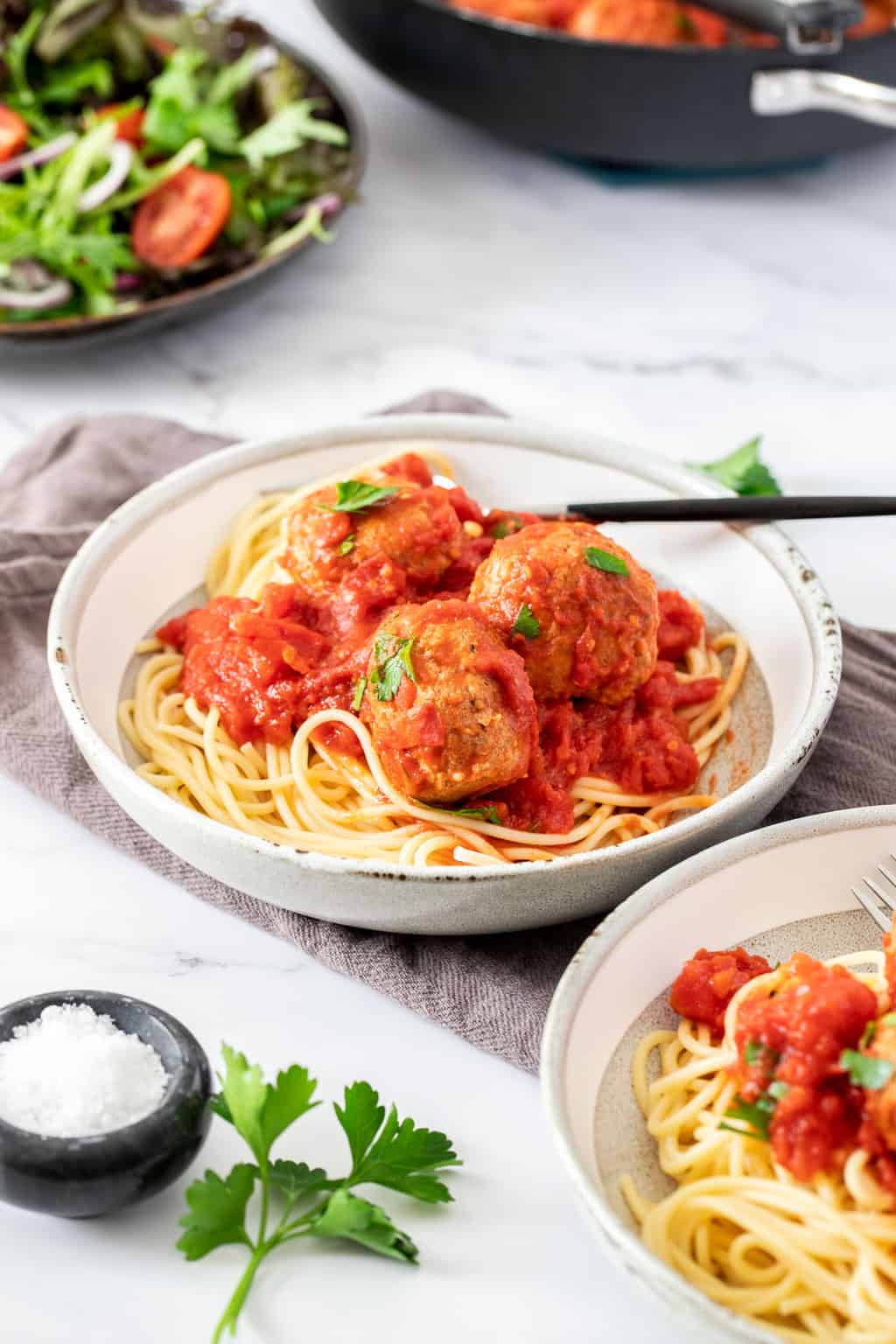 Two bowls of spaghetti with healthy turkey meatballs on top, In the back is a small plate of salad and the skillet of the rest of the meatballs