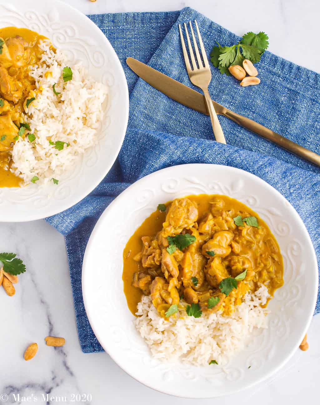 An overhead shot of 2 bowls of peanut butter chicken curry surrounded by peanuts, cilantro, and a blue towel