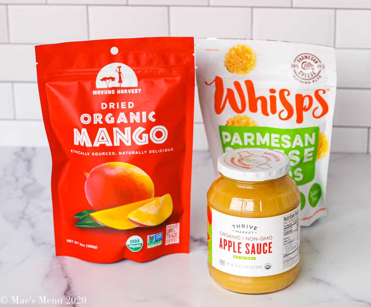 Snacks from thrive market on the counter: a bag of mangoes, a bottle of applesauce, and a bag of parmesan cheese whisps