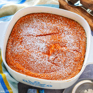 A large white dish of carrots souffle surrounded by blue towels, a wooden spoon holder, and a shaker of confectioner's sugar