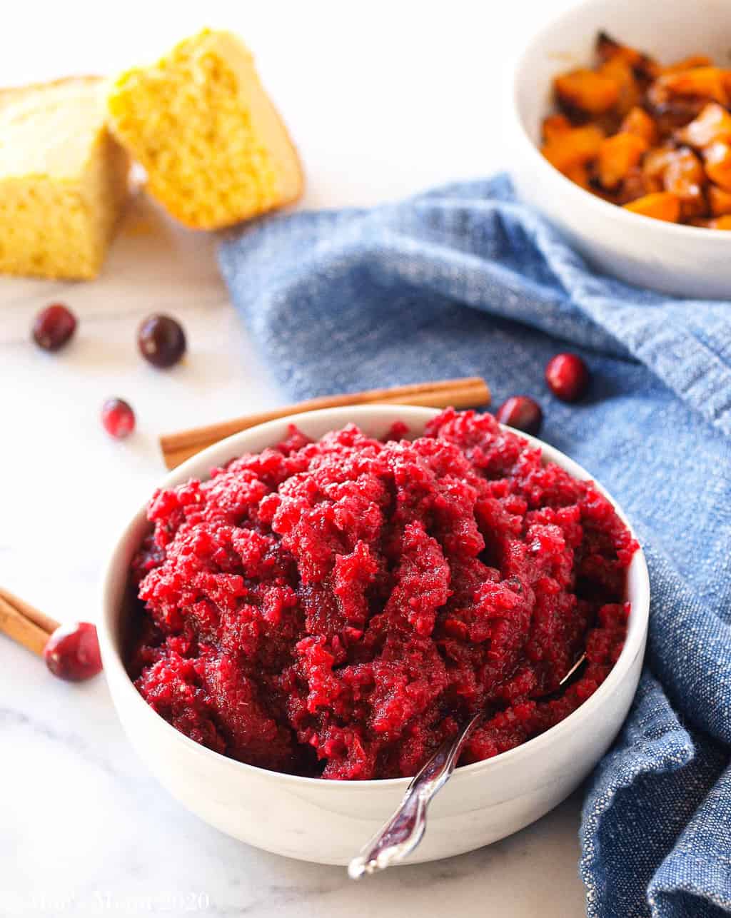 A bowl of apple cranberry sauce on a granite counter with cornbread, air fryer butternut squash, and a blue towel