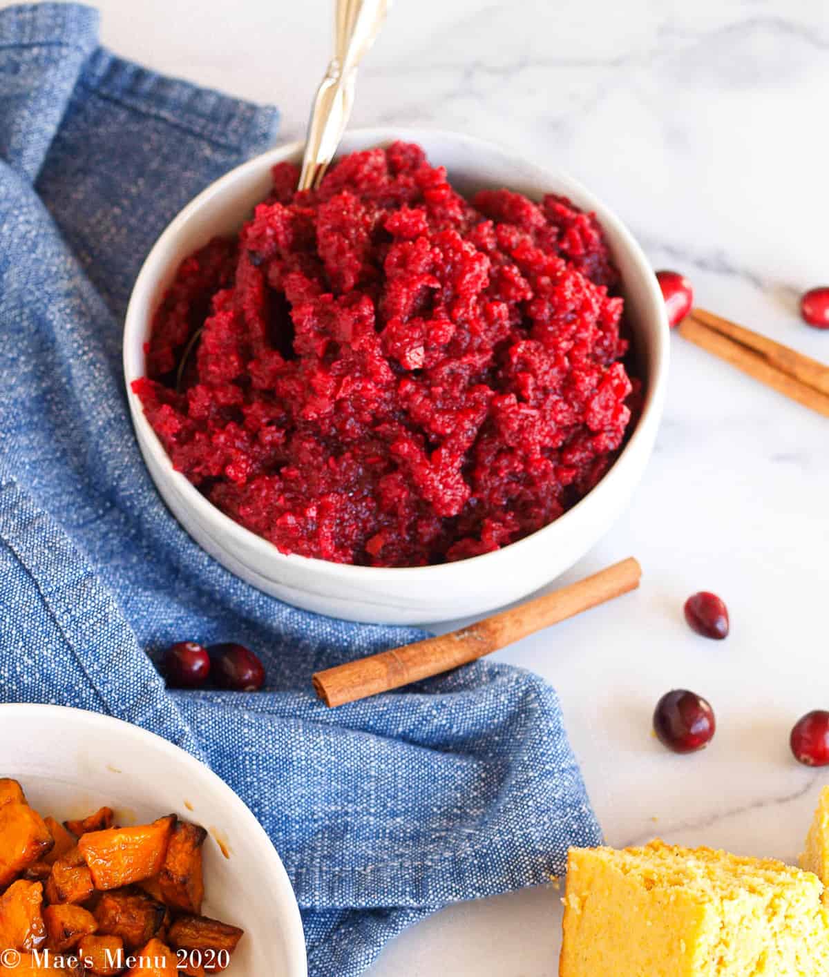 A bowl of apple cranberry sauce on a counter next to a blue towel, cinnamon sticks, and cranberries