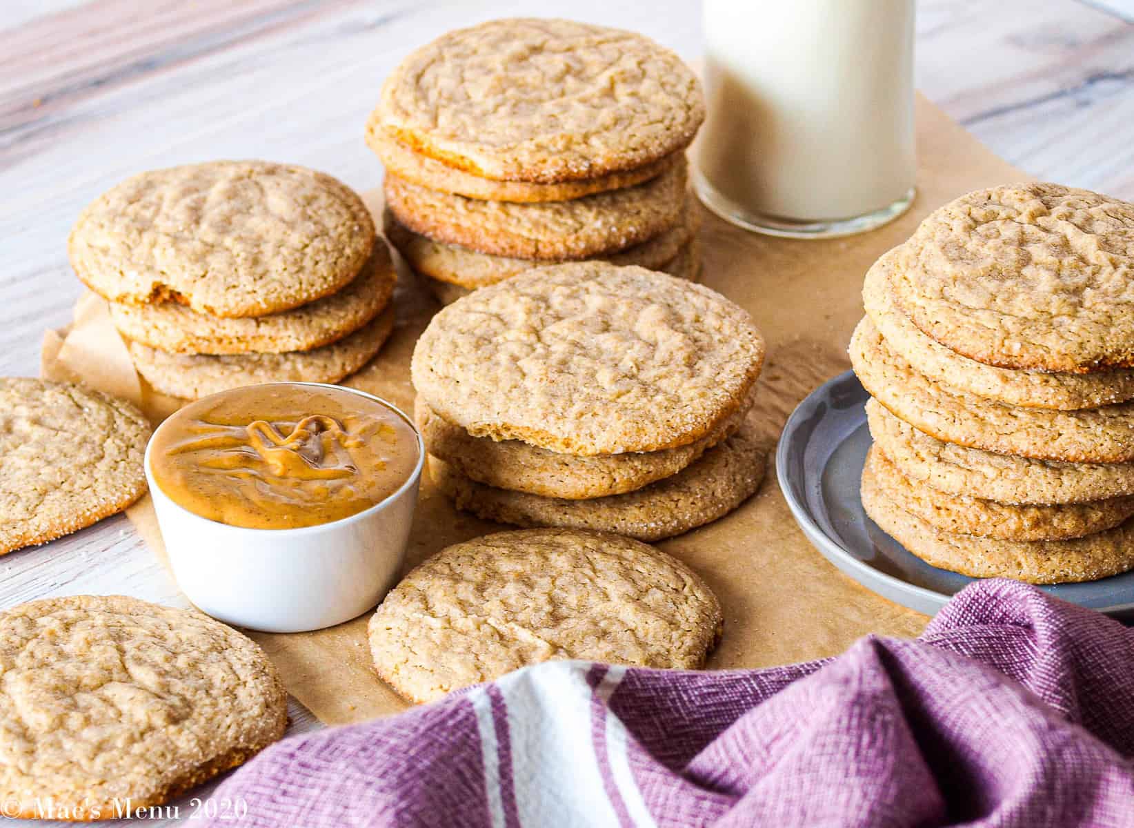 A side angle shot of stacks of whole wheat peanut butter cookies on a sheet of parchment paper with a cup of peanut butter and a glass of milk