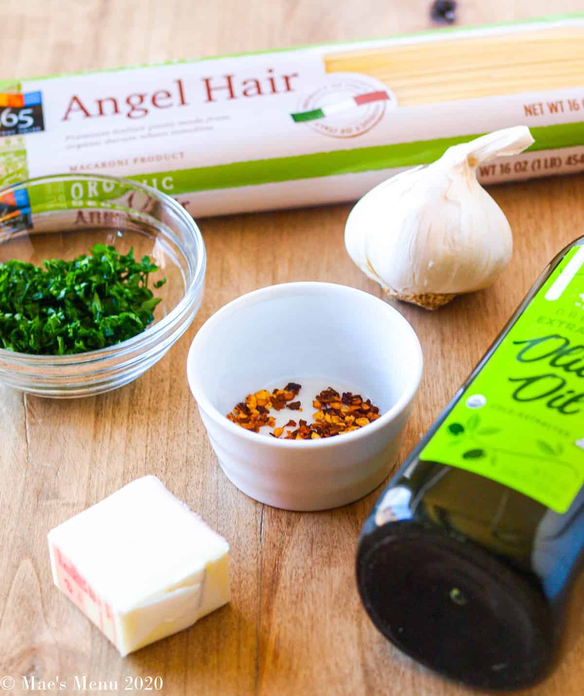 all the ingredients for garlic angel hair pasta on a wooden table: angel hair, chopped parsley, garlic, red pepper flakes, olive oil, and butter