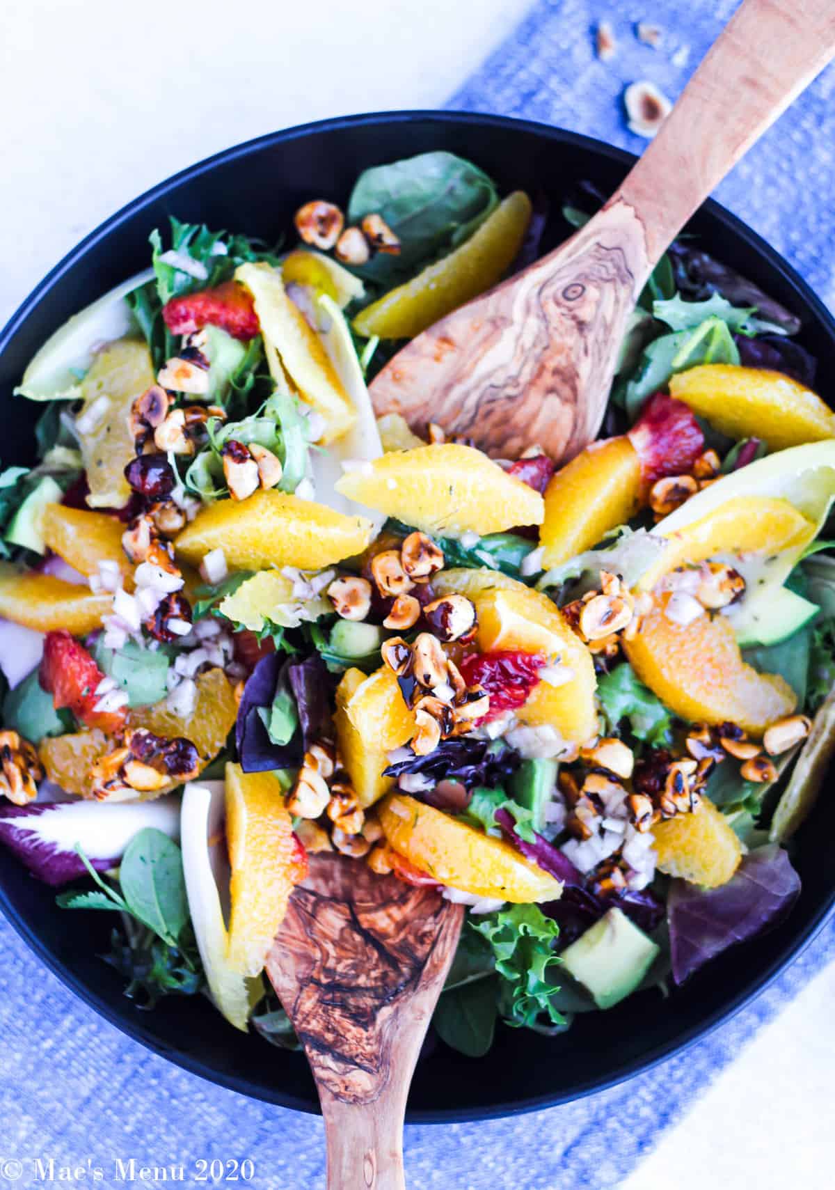 An overhead shot of a black bowl full of orange salad with honeyed hazelnuts with salad tongs in the salad