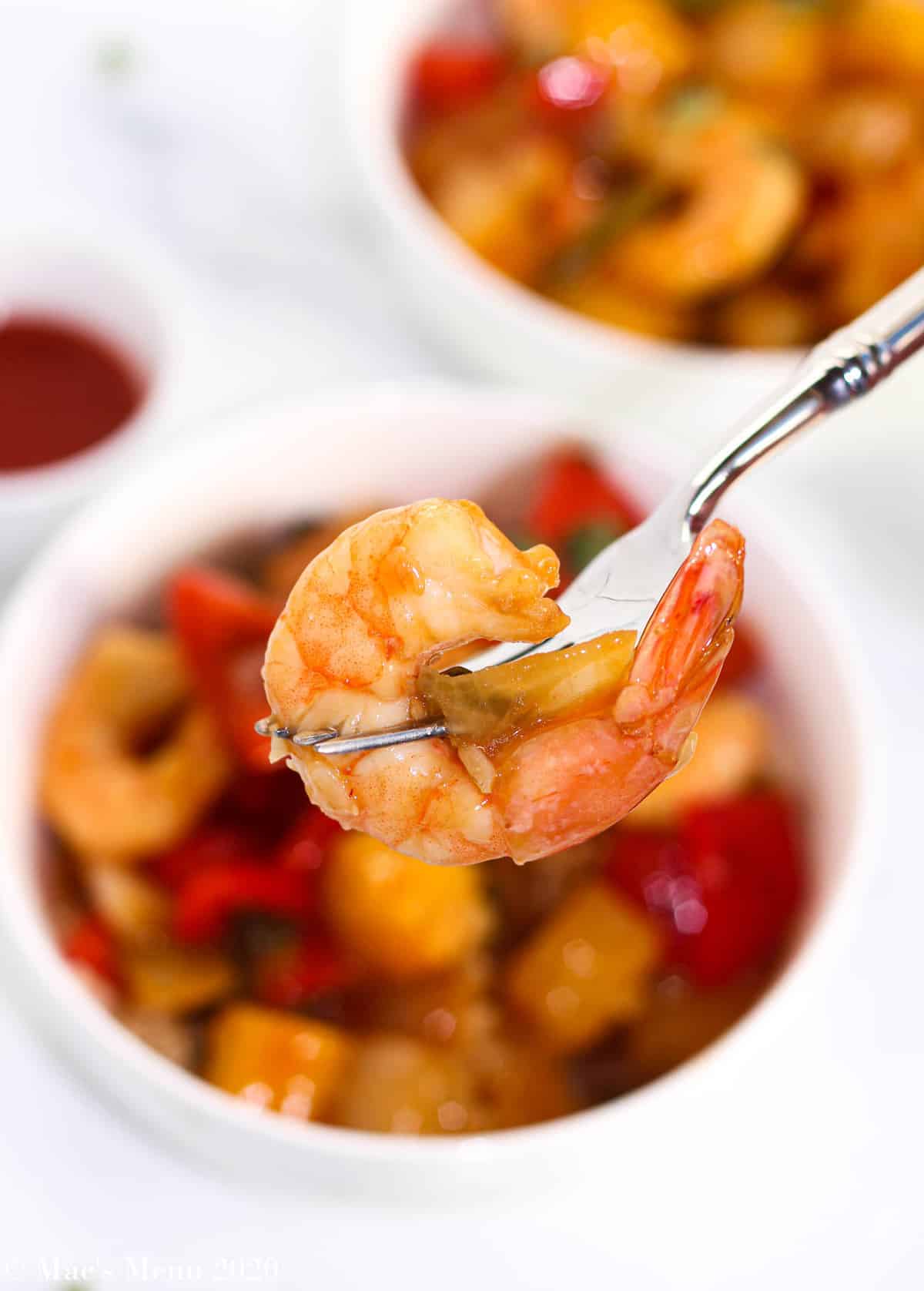 An up-close shot of sweet and sour shrimp on a fork with the bowls of shrimp in the background