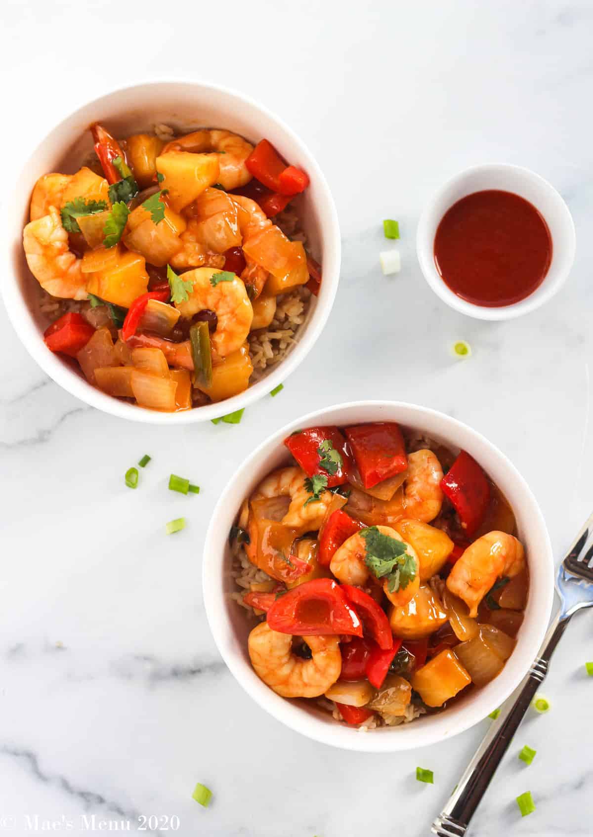 An overhead shot of two bowls of sweet and sour shrimp next to a cup of sriracha and sliced green onions
