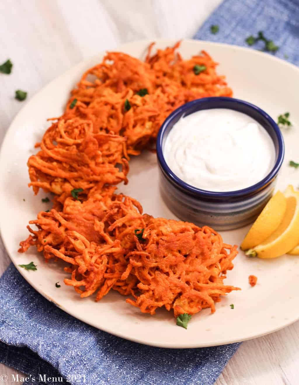 An angled shot of spiced sweet potato fritters on a white plate with a lemon sour cream dipping sauce and lemon slices