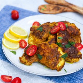 A plate of rubbed turmeric chicken on a white plate with lemons, tomatoes, and cilantro