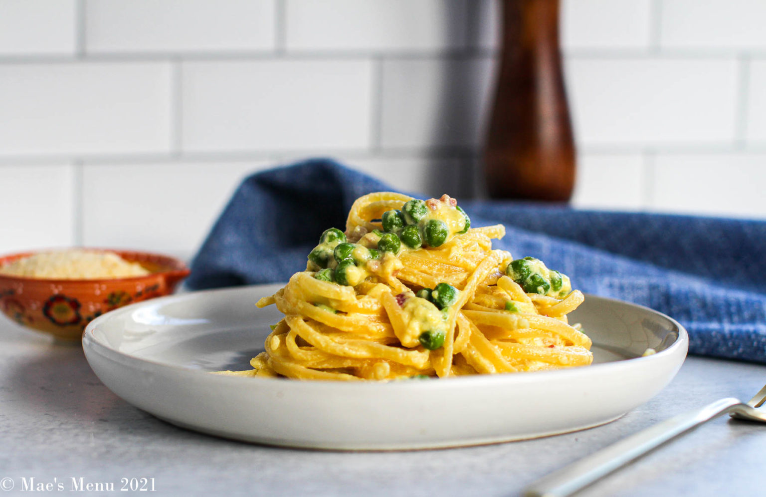 A plate of fettuccine pasta with peas and a creamy sauce is placed on a white plate. A small bowl of grated cheese and a fork are on the table beside it. A blue cloth and a peppermill are in the background, with a tile backsplash behind them.