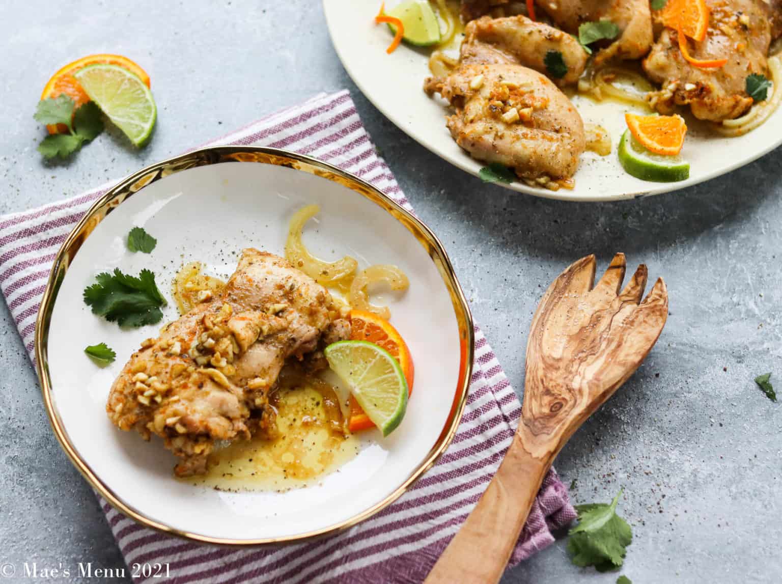 A plate of citrus-glazed chicken pieces garnished with lime, orange slices, and cilantro. The dish is presented on a white plate with a gold rim, placed on a striped napkin. A wooden serving fork is beside the plate; more chicken is on a large serving dish in the background.