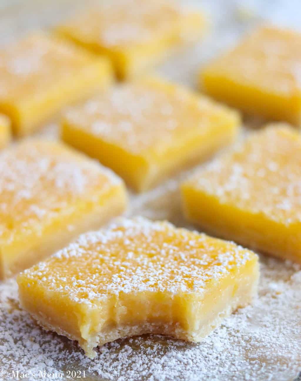 Lemon bars on parchment paper with the first one with a bite taken out of it 