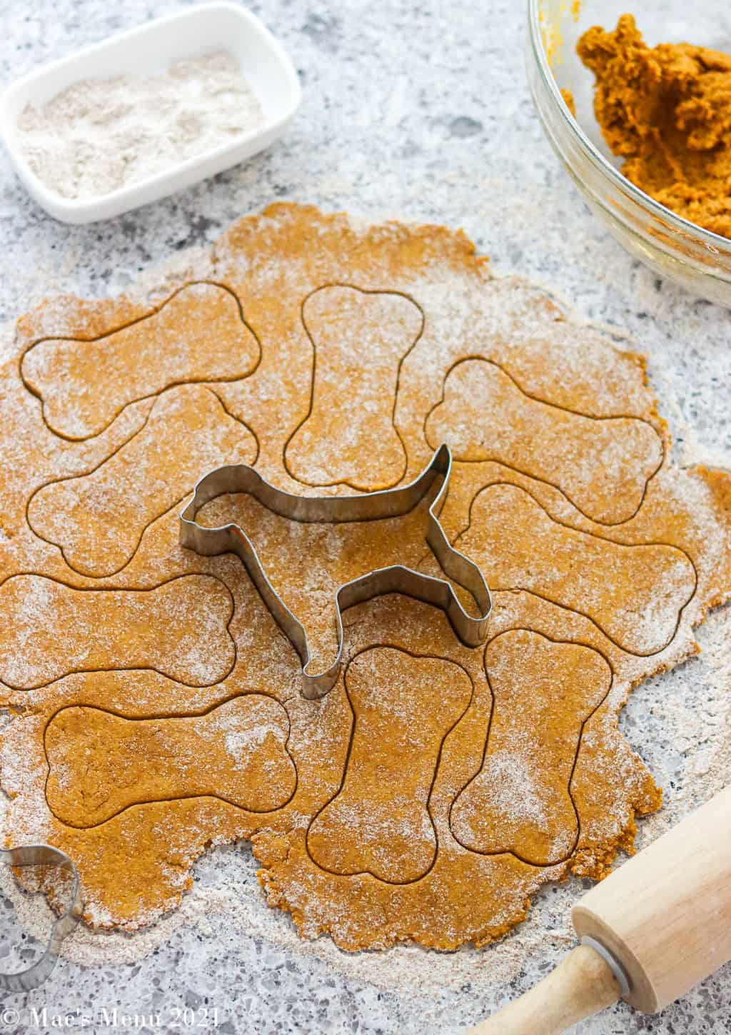 An overhead shot of dog treat dough rolled out with cookie cutter shapes cut out and a dog shape cutter in the center of the dough