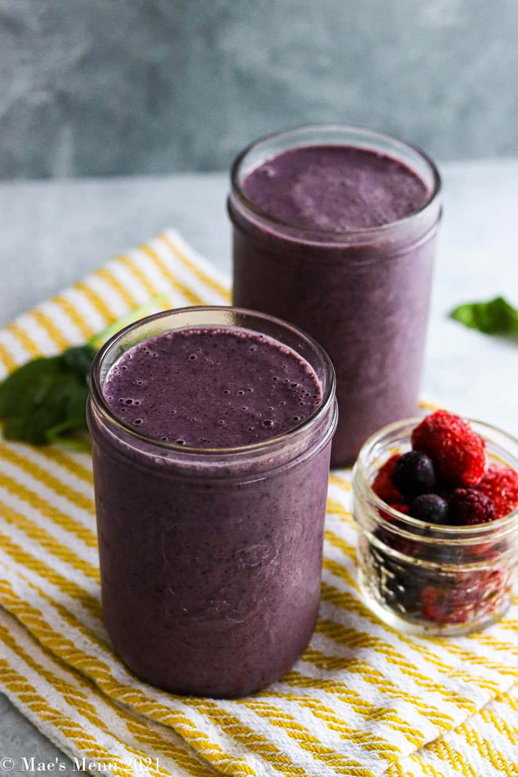 Two glasses of blueberry spinach smoothie on a gold striped towel next to a cup of frozen berries