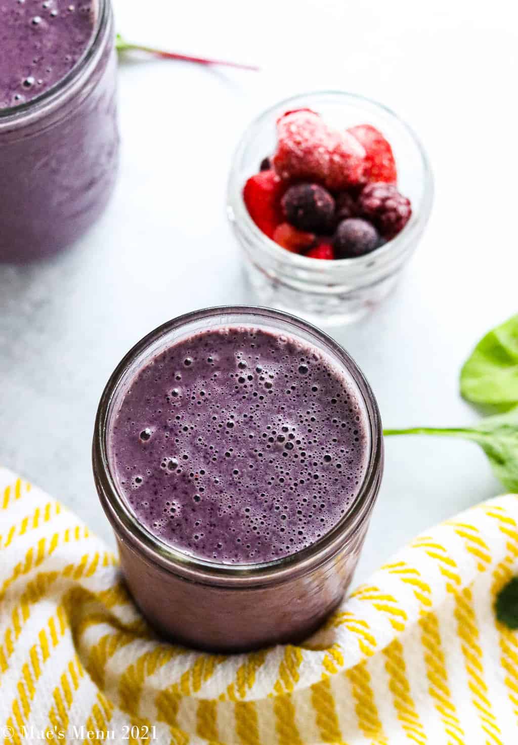 An up-close overhead shot of a cup of blueberry spinach smoothie with a cup of frozen berries in the background