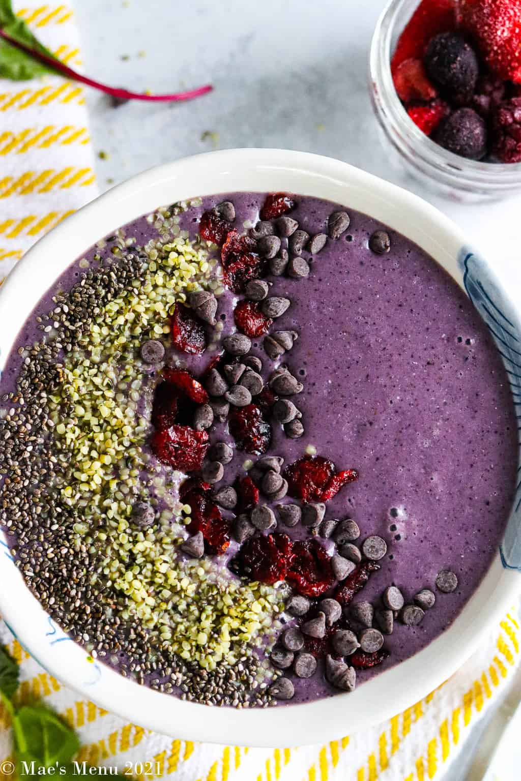 An up-close overhead shot of a spinach and blueberry smoothie bowl with chia seeds, hemp seeds, cranberries, and chocolate chips