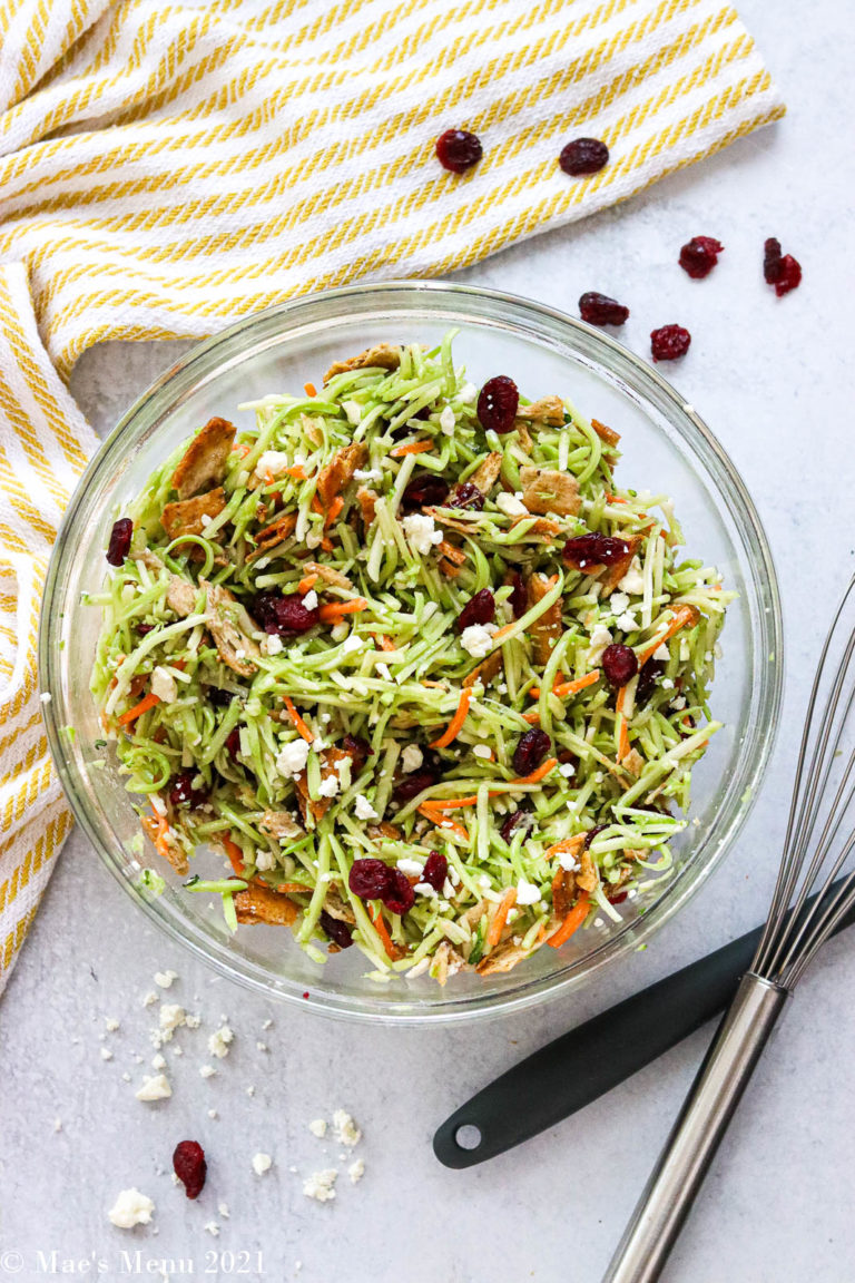 A large glass mixing bowl of broccoli slaw next to a rubber scraper and whisk