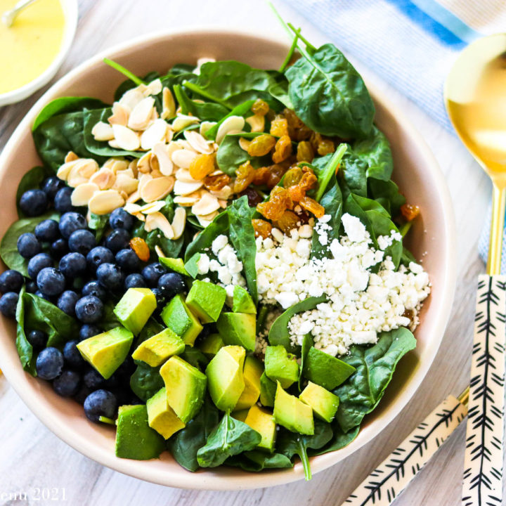 A horizontal shot of a large bowl of blueberry avocado salad surrounded by a dish of dressing and salad tongs