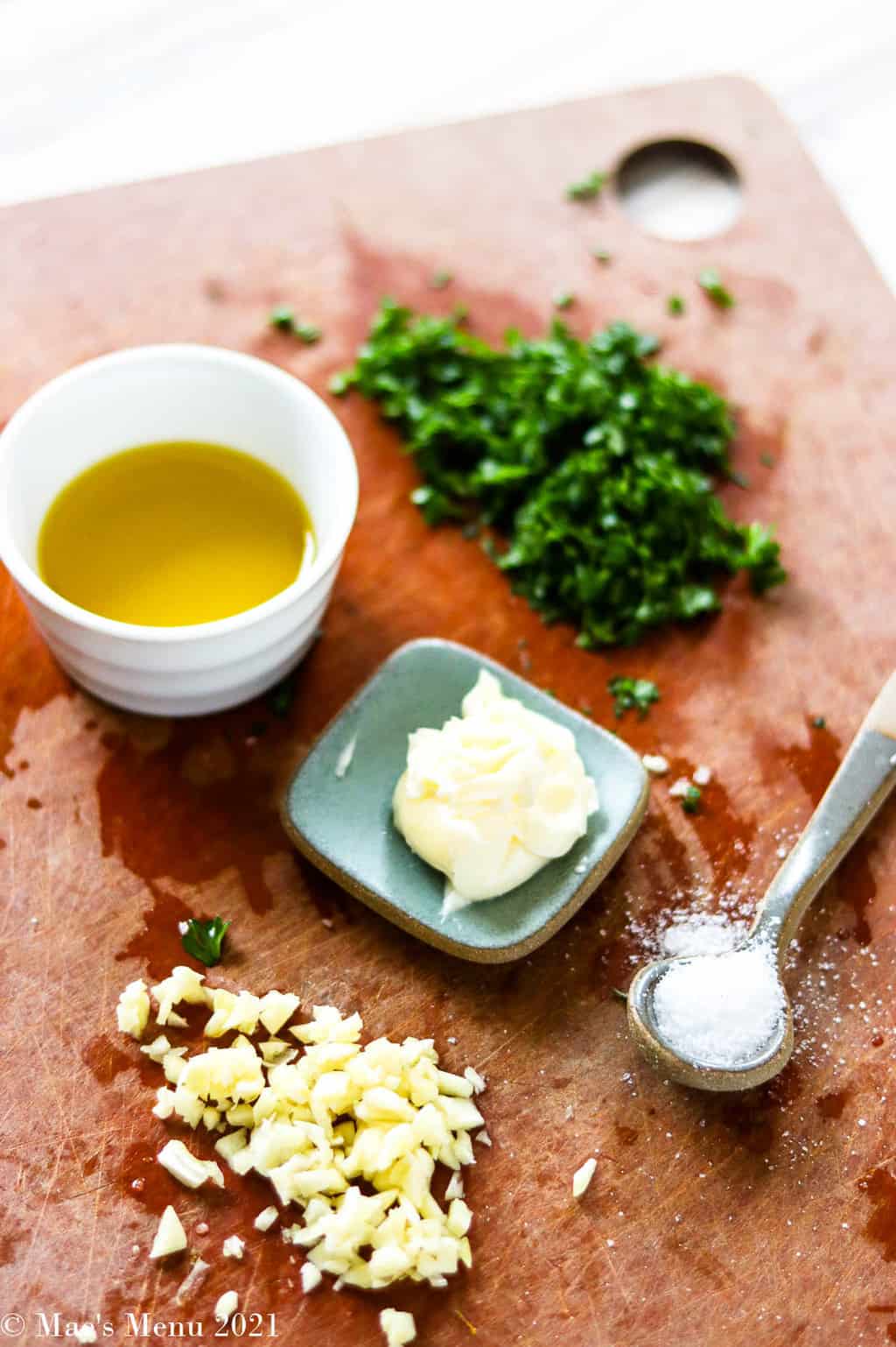 A cutting board with parsley, butter, salt, olive oil, and garlic on it