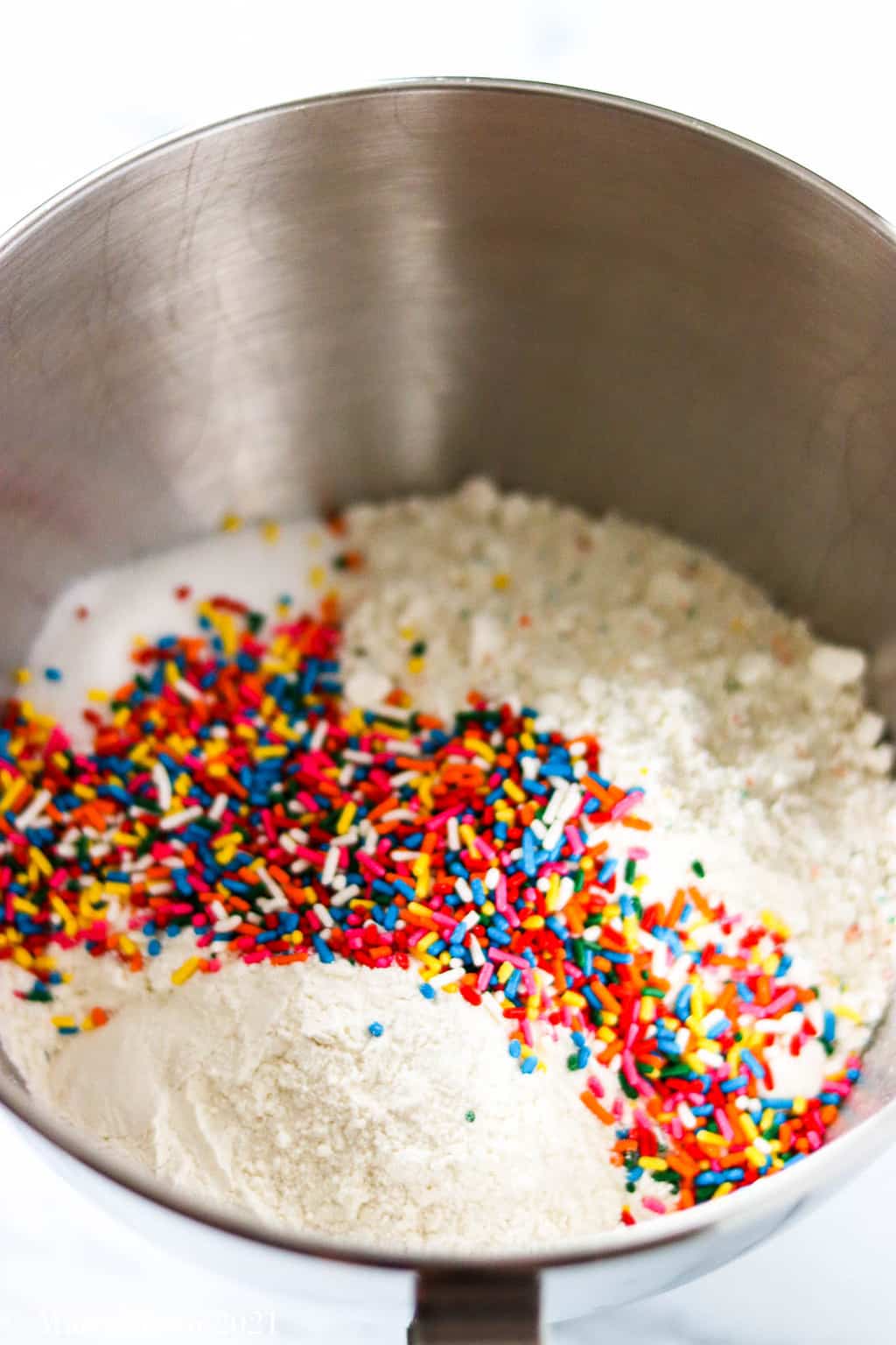 A mixing bowl with confetti cake mix, flour, sugar, and sprinkles