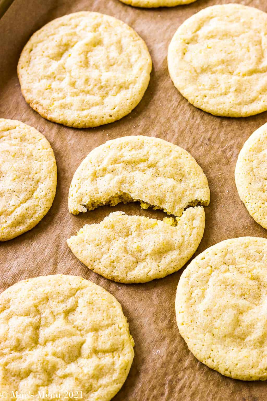 A lemon sugar cookie broken in half on a baking tray of lemon sugar cookies