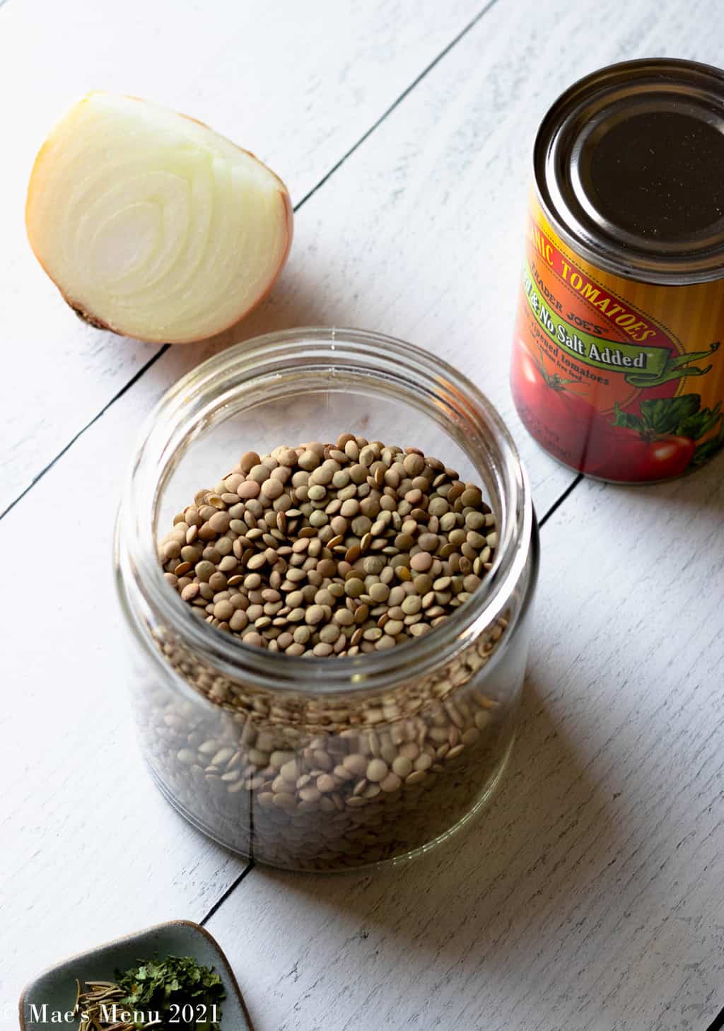 A clear glass container of lentils surrounded by half an onion, a can of tomatoes, and herbs