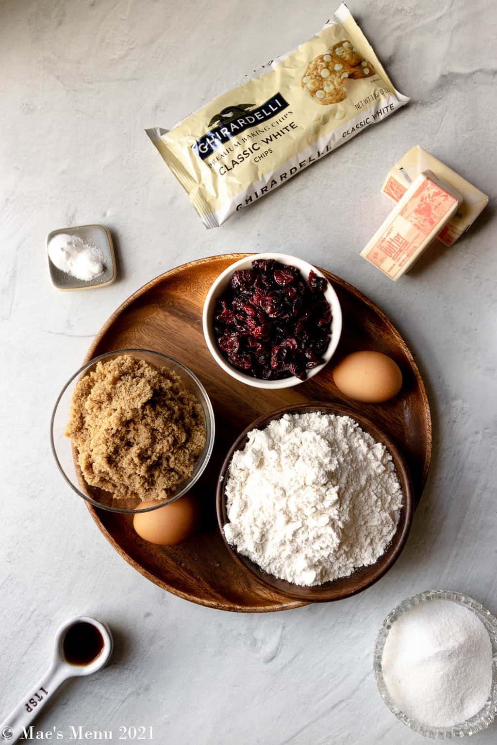 All the ingredients for brown butter white chocolate chip cookies: white chocolate chips, baking soda, butter, dried cranberries, brown sugar, white sugar, flour, eggs, and vanilla extract