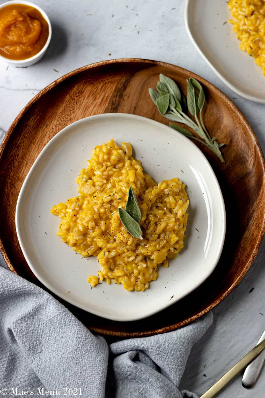 A serving of pumpkin risotto on a dish surrounded by herbs, silverware, and a small dish of pumpkin