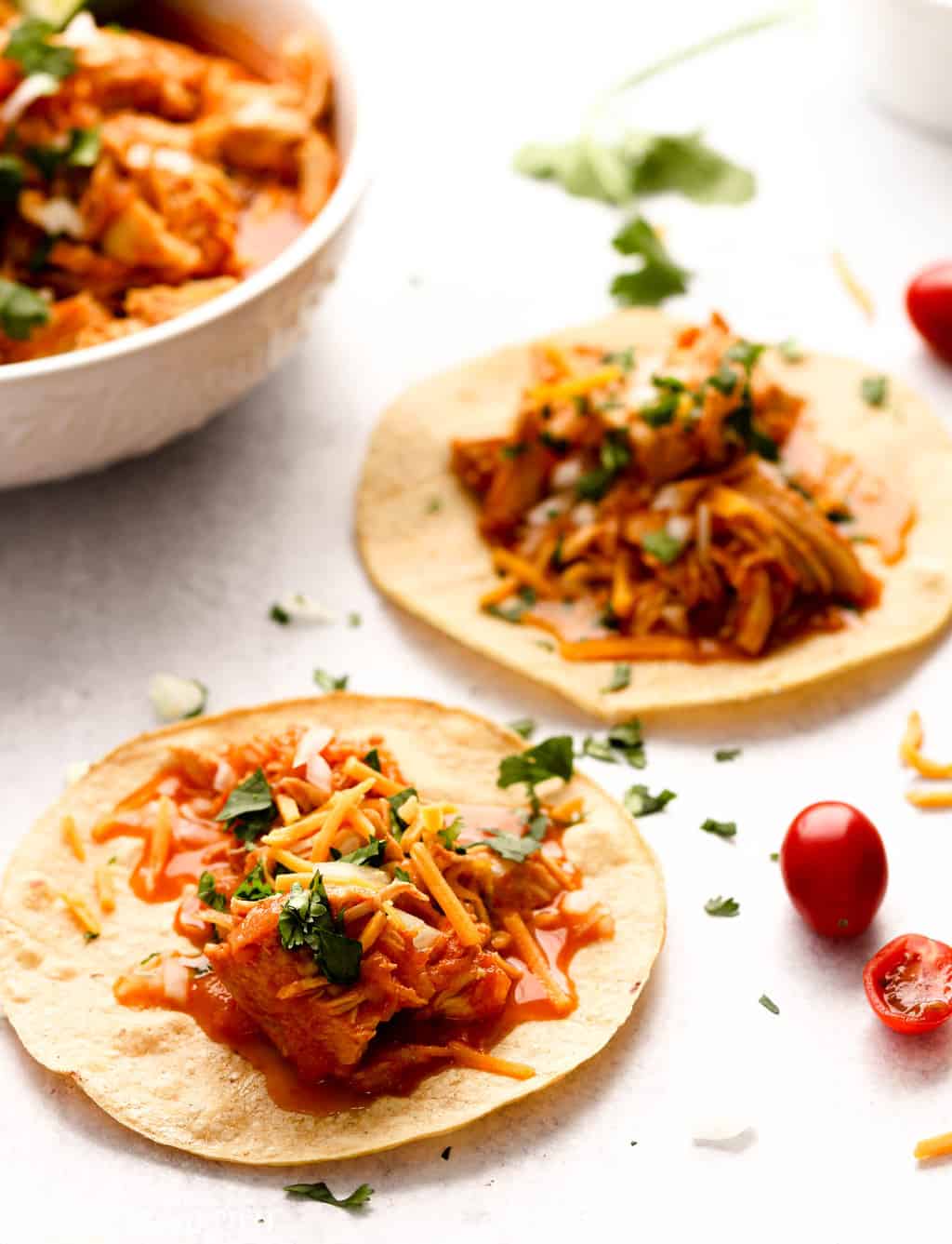Two instant pot chicken tacos on a concrete counter in front of a bowl of chicken taco meat