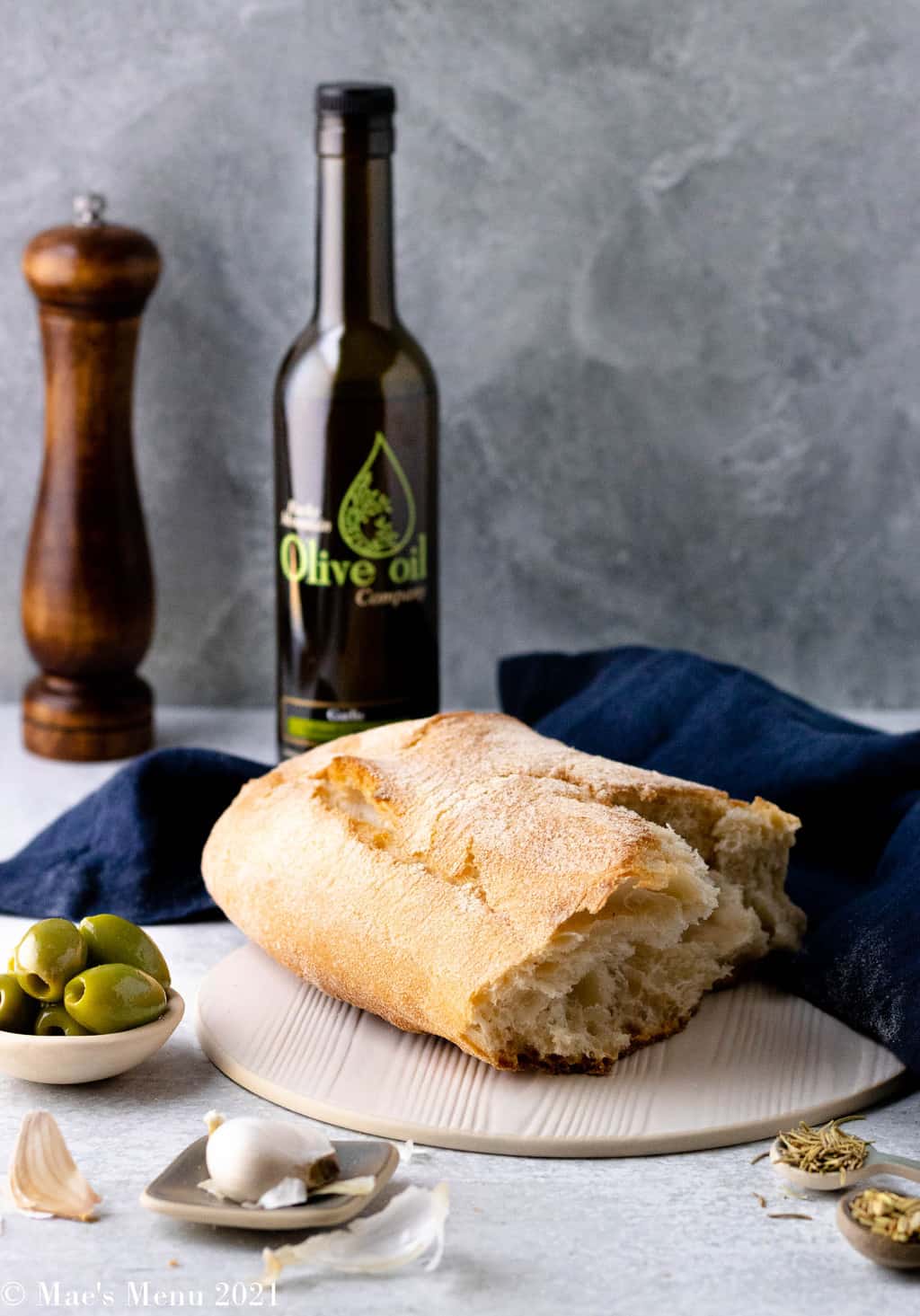 A torn loaf of ciabatta bread with a bottle of olive oil, olives, and seasonings