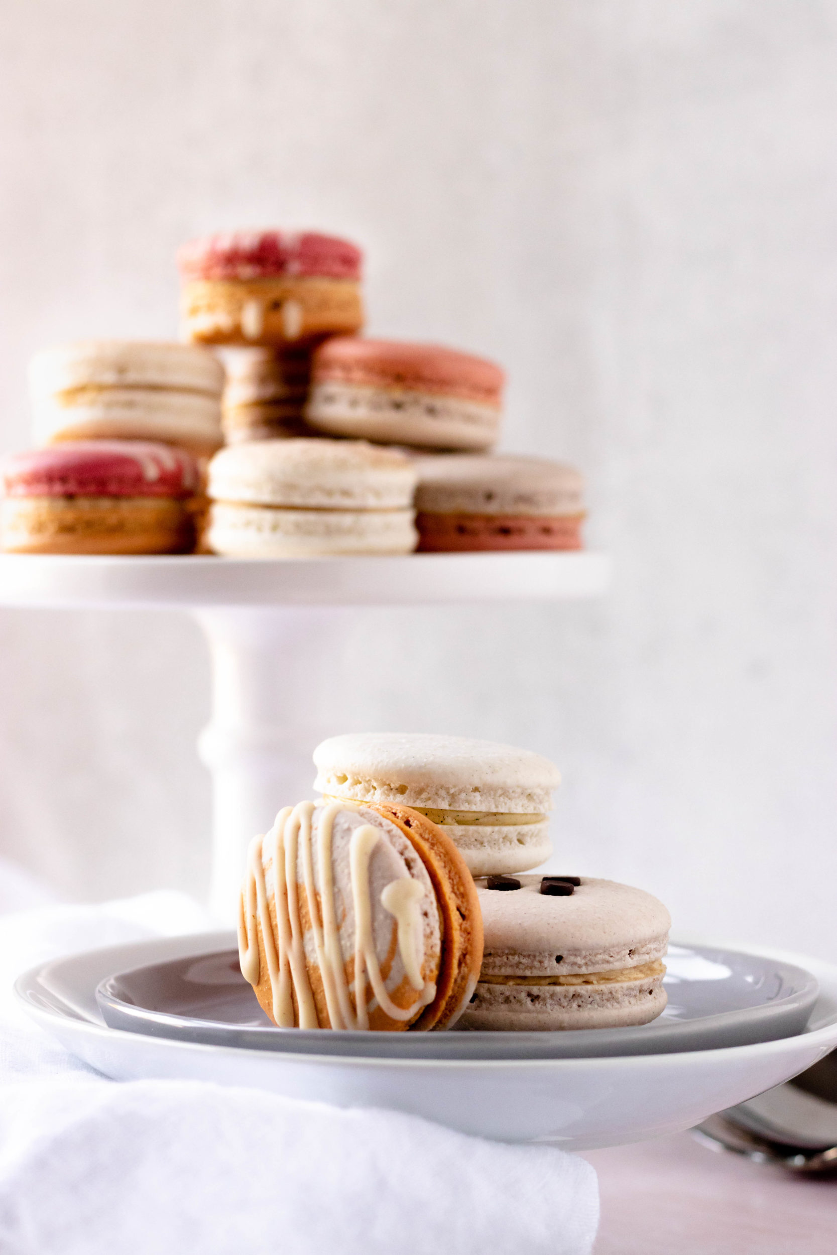 A cake platter of macarons in the background with layered plates with macarons on them in the foreground.